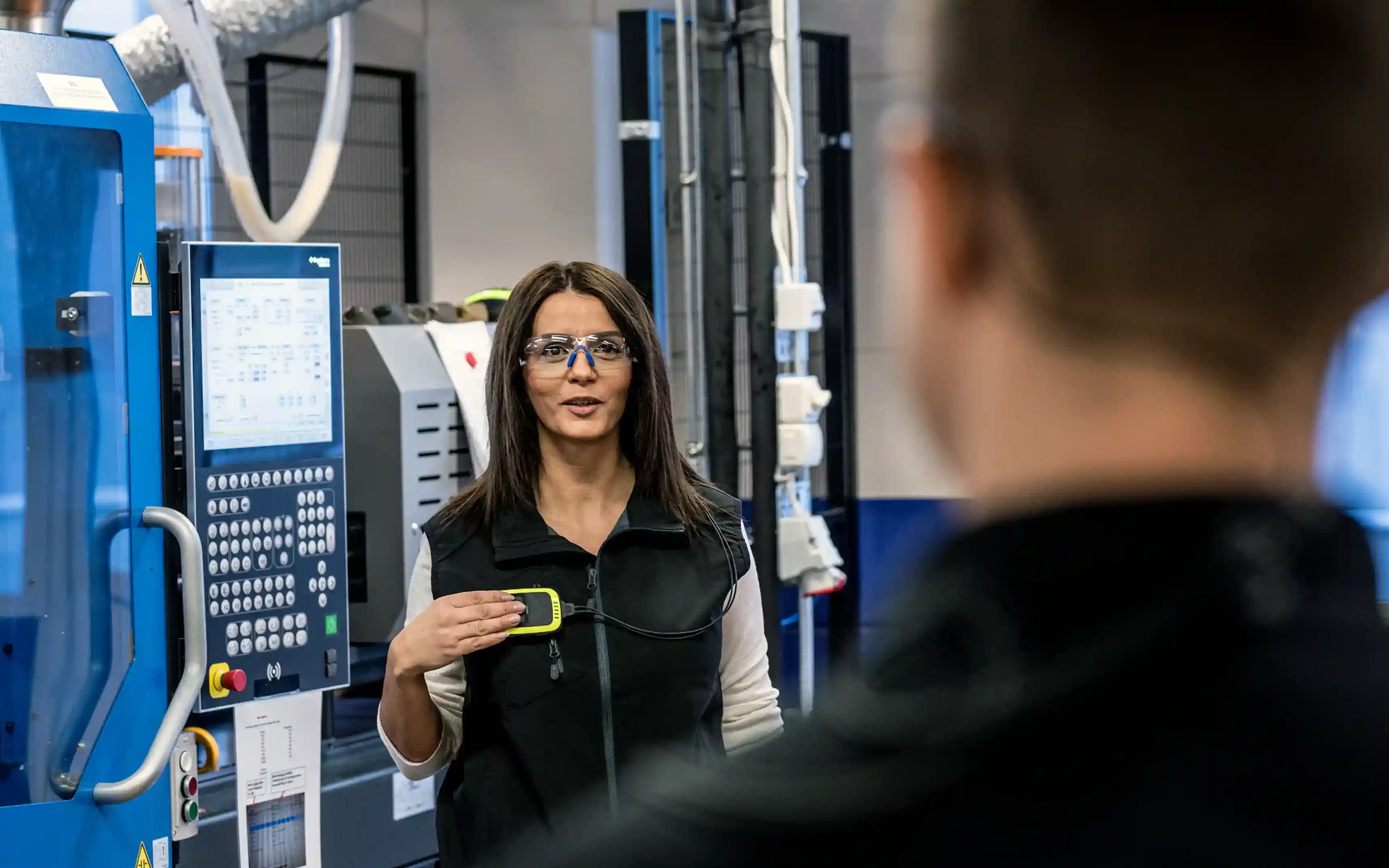 En medarbetare klädd i svart väst och skyddsglasögon står vid en CNC-maskin och genomför ett anrop på en 3M Peltor PIC-100 trådlös intercom som hänger på bröstet. I förgrunden syns en hennes kollega ur fokus.