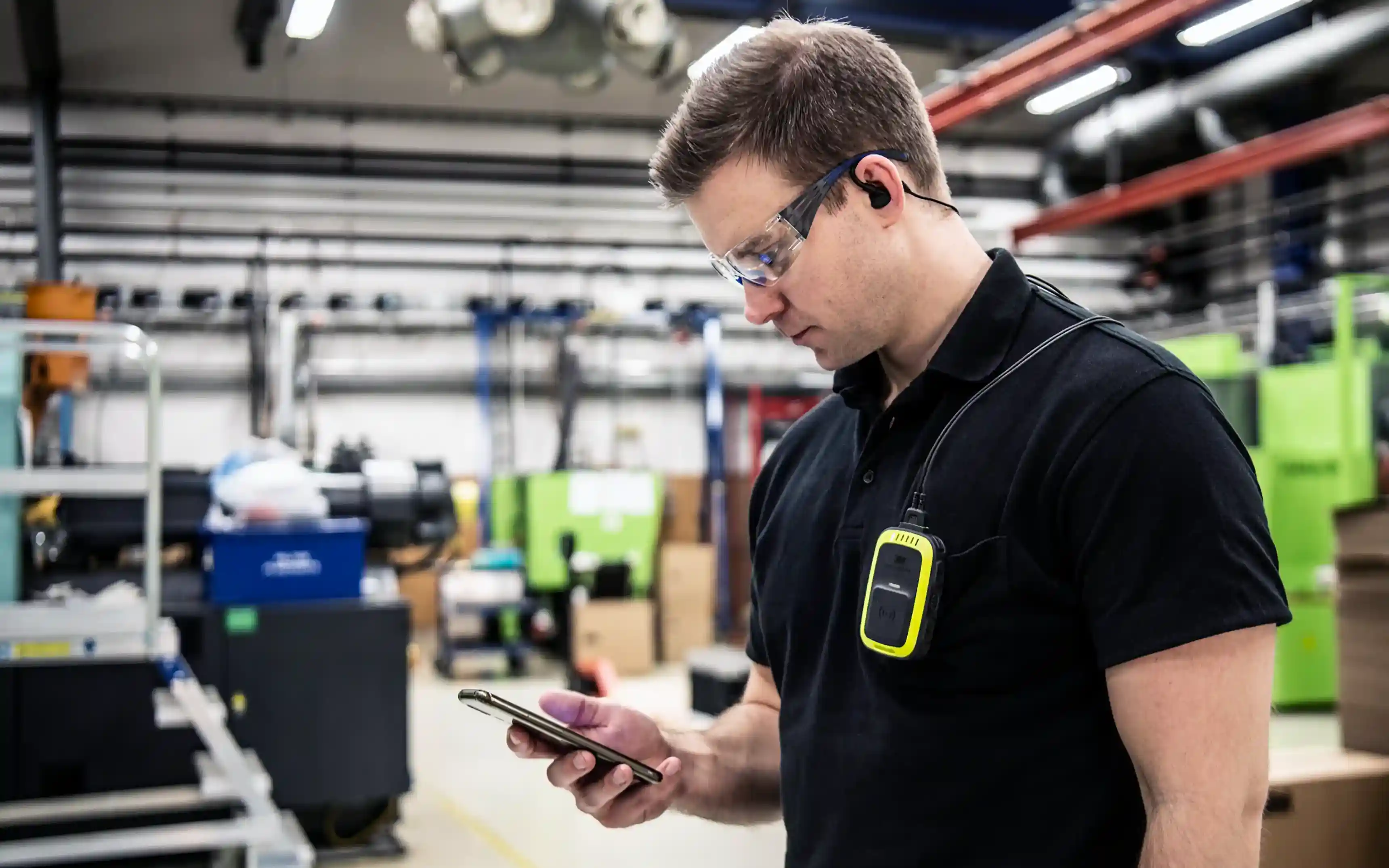 En medarbetare klädd i svart med skyddsglasögon som står i en produktionslokal och styr sin 3M Peltor PIC-100 trådlös intercom med mobiltelefonen.