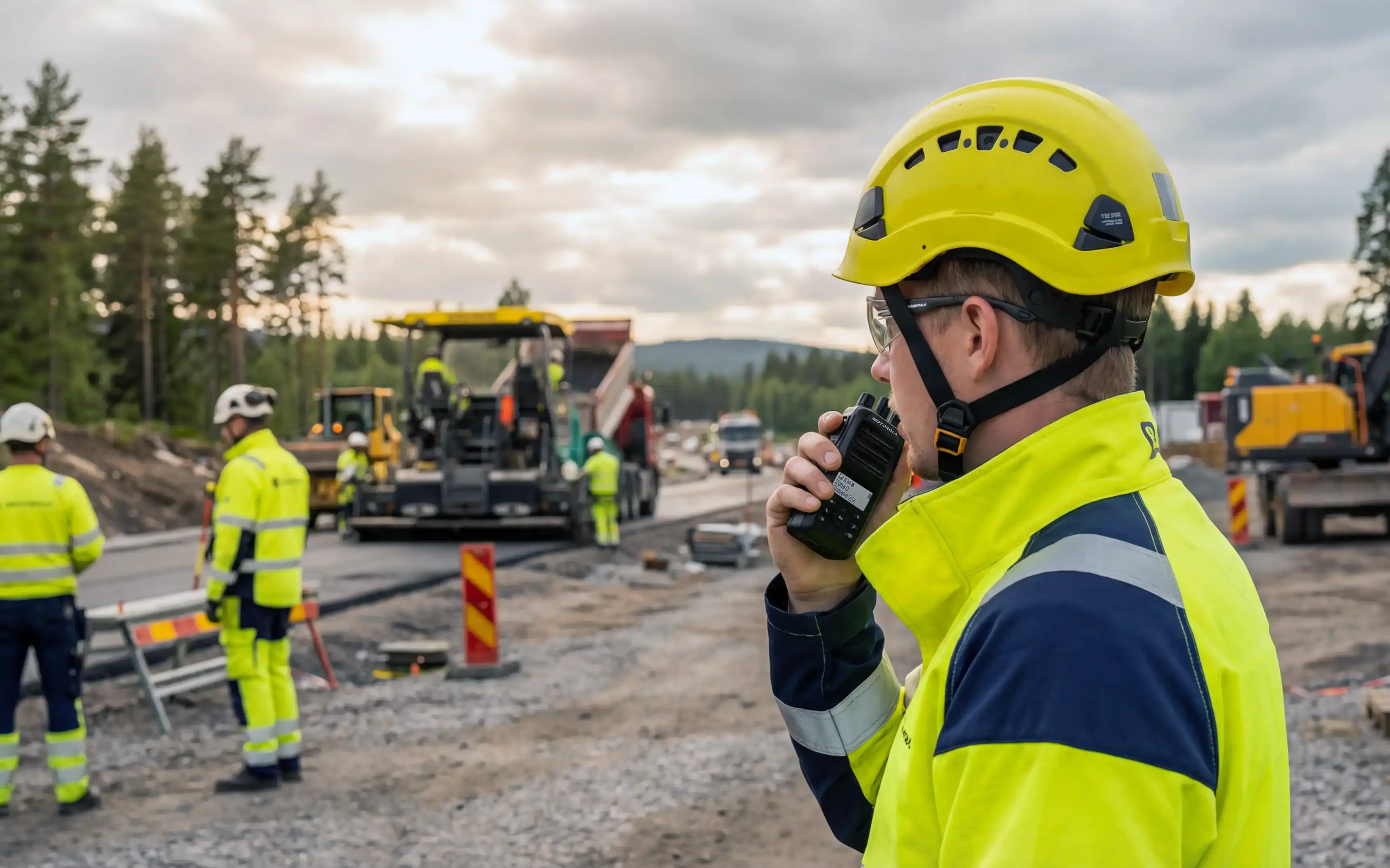 En person som står ute på ett vägarbete i pratar i en komradio med asfaltsläggning som pågår i bakgrunden.