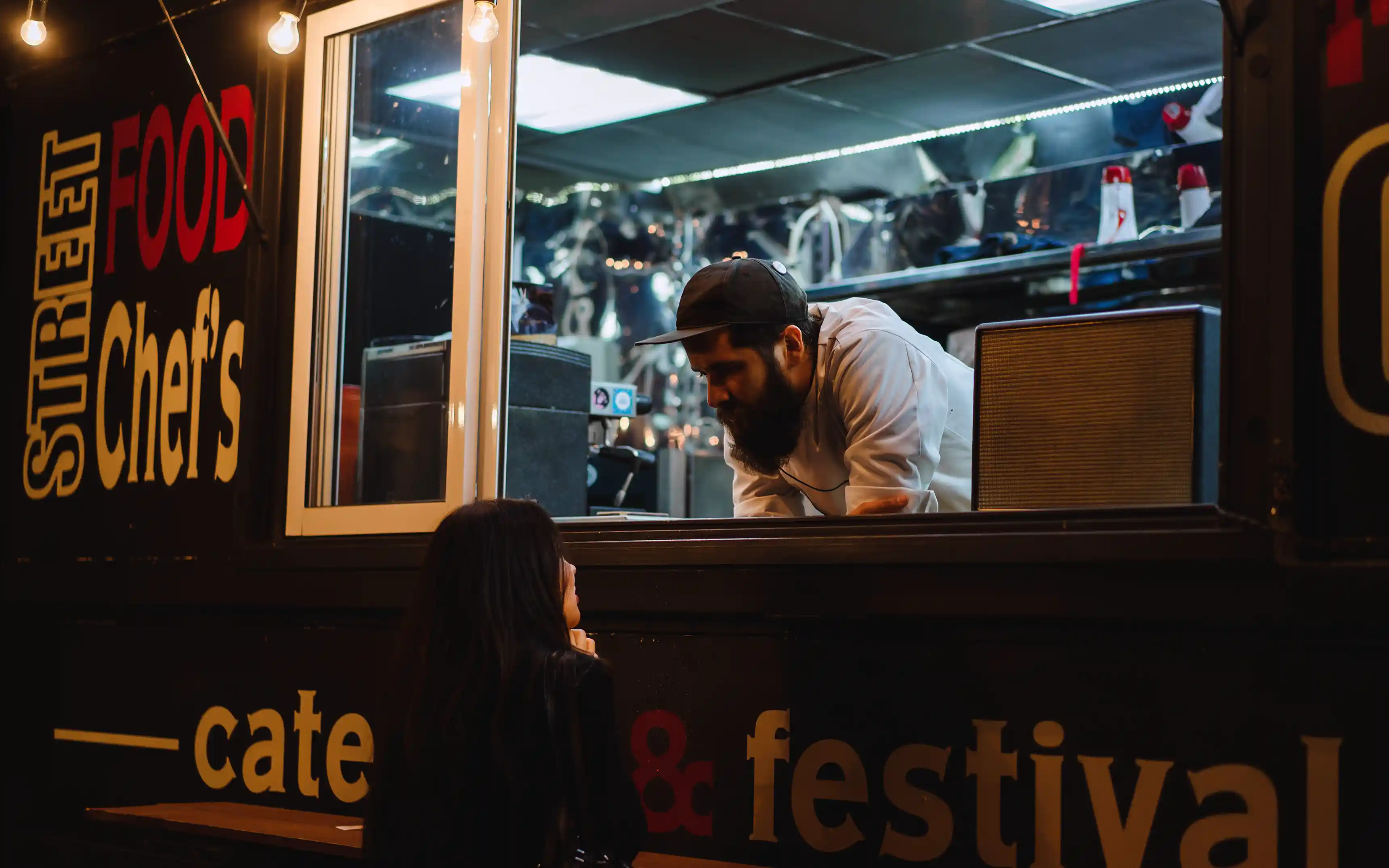 En man som står inne i en svart food truck och pratar med en kund som står utanför.