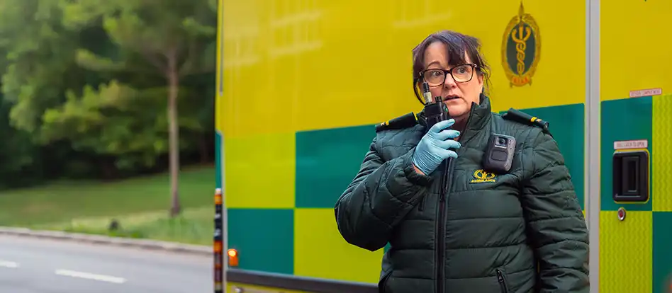 En ambulanssjukvårdare som gör ett radioanrop. I bakgrunden ambulansen och en del av en väg och några träd.