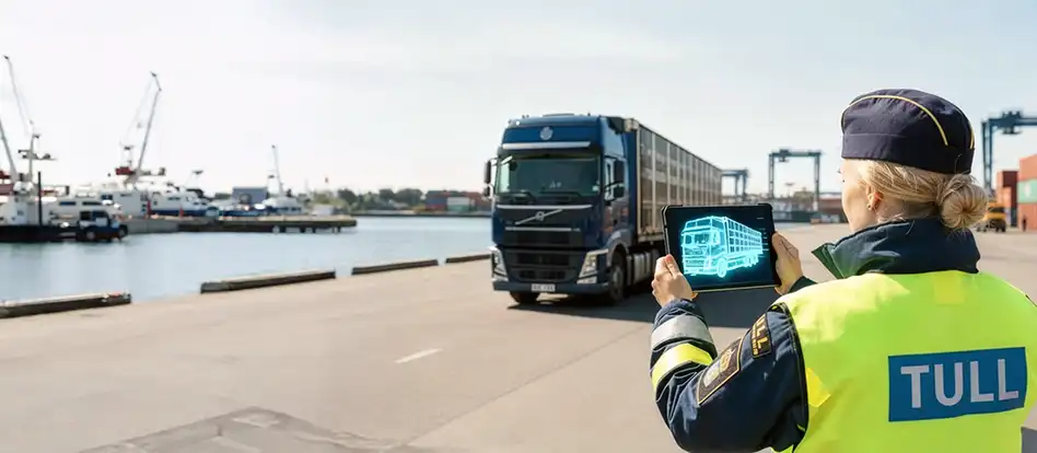En tulltjänsteman som tittar på en surfplatta som visar en scan av lastbilen som kör förbi. I bakgrunden syns en hamnmiljö med kranar, containrar och en kaj.
