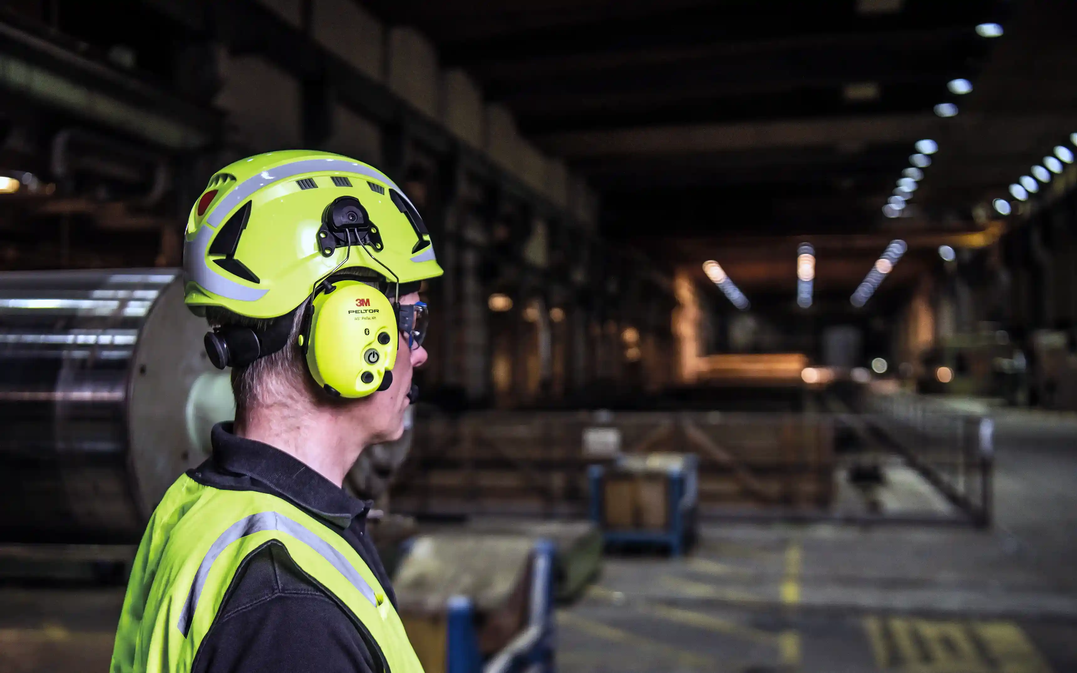 Industriarbetare i varselkläder som bär ett bullerdämpande kommunikationsheadset monterad på sin hjälm inne i en stor industrilokal.